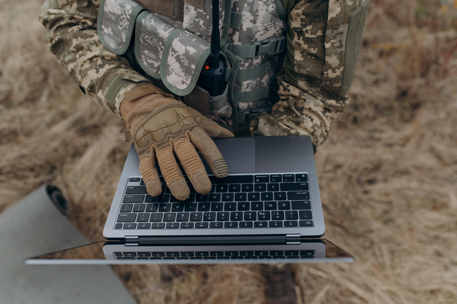 A person in field clothing using a laptop, with a radio carried on their vest.